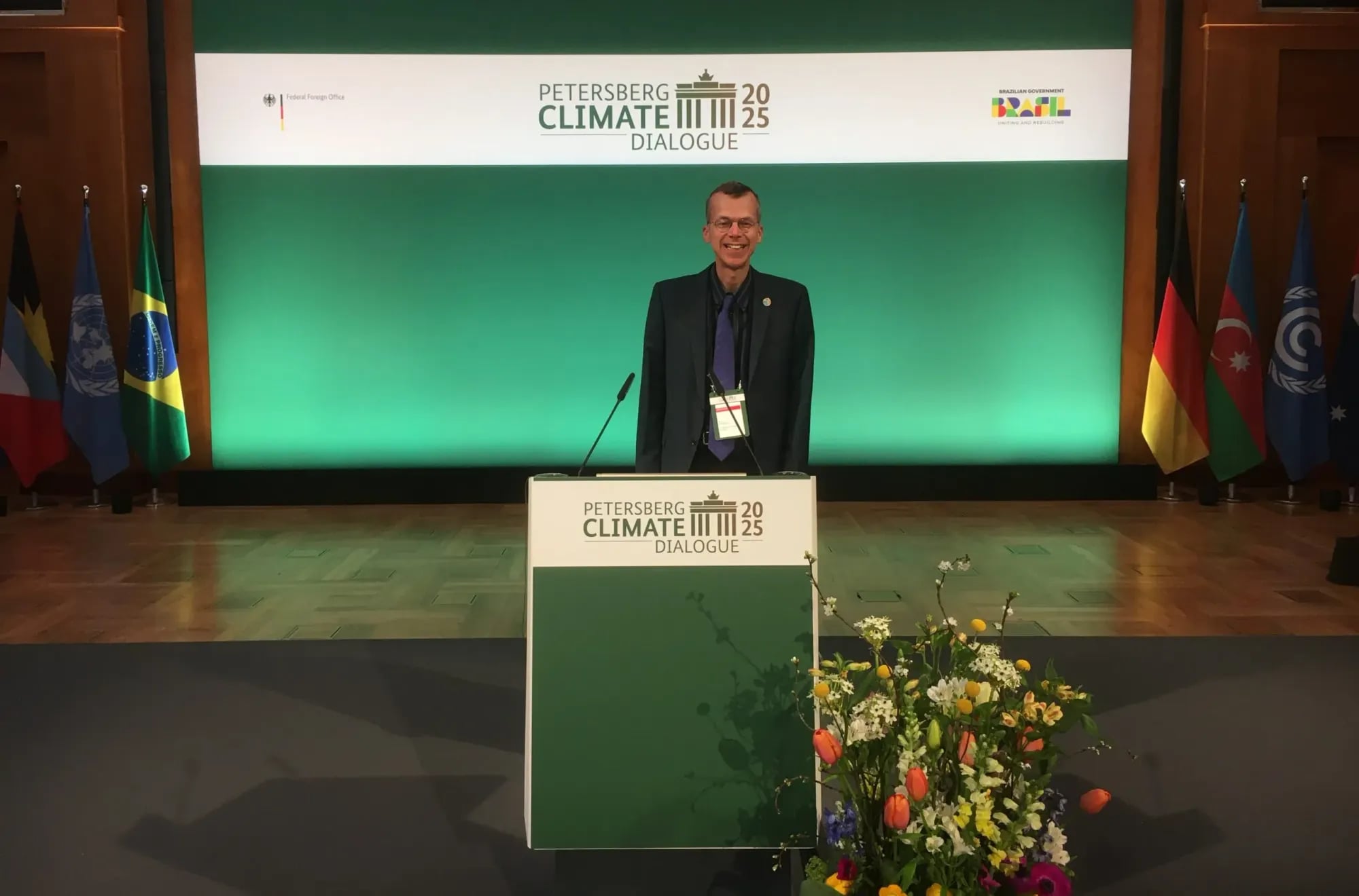 A speaker at the podium of the Petersberg Climate Dialogue 2025 in Berlin, flanked by rows of national flags